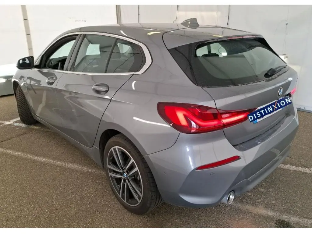 BMW Série 1 116d Business Design gris vue 3/4 arrière droit, feux arrière allumés et jantes alliage visibles.