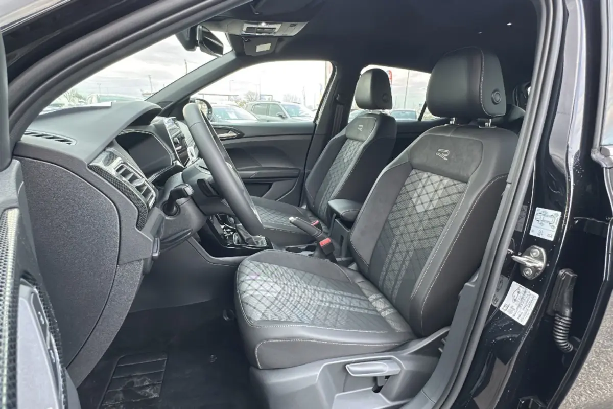 Vue intérieure côté conducteur d'un Volkswagen T-Cross noir intense 2025, sièges tissu R-Line avec surpiqûres blanches.