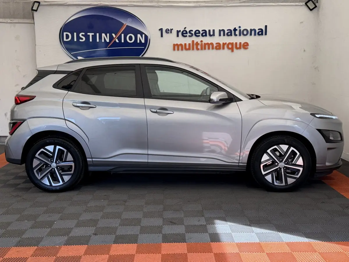 Vue de profil côté gauche d'un Hyundai Kona électrique gris clair garé en intérieur avec logo Distinxion en arrière-plan.