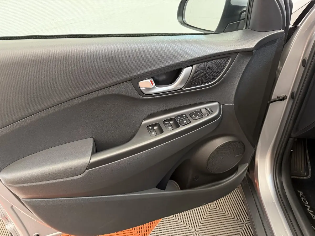 Vue intérieure côté gauche de la porte avant gris clair du Hyundai Kona électrique avec commandes de vitres et haut-parleur intégré.