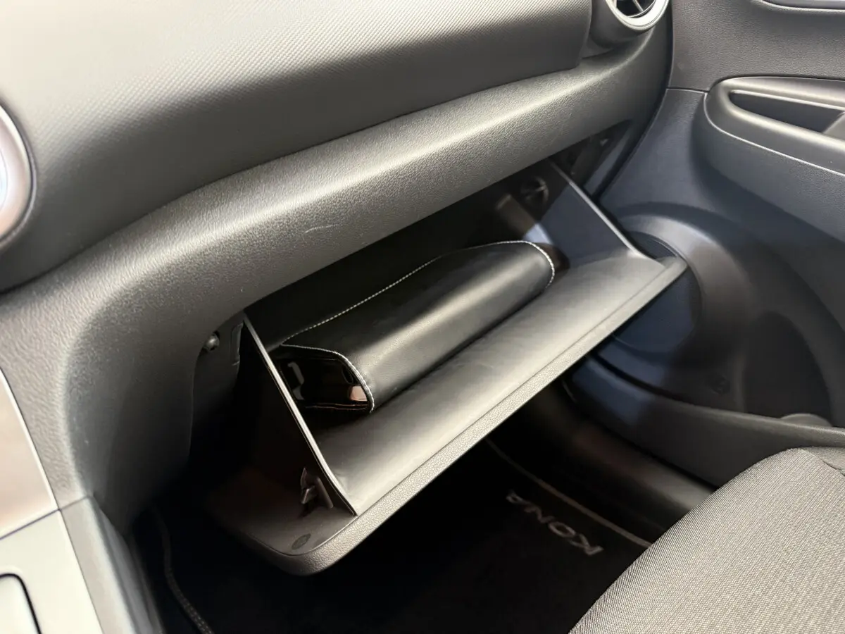 Intérieur du Hyundai Kona électrique 2022, vue sur la boîte à gants ouverte avec pochette noire et tapis de sol marqué Kona.