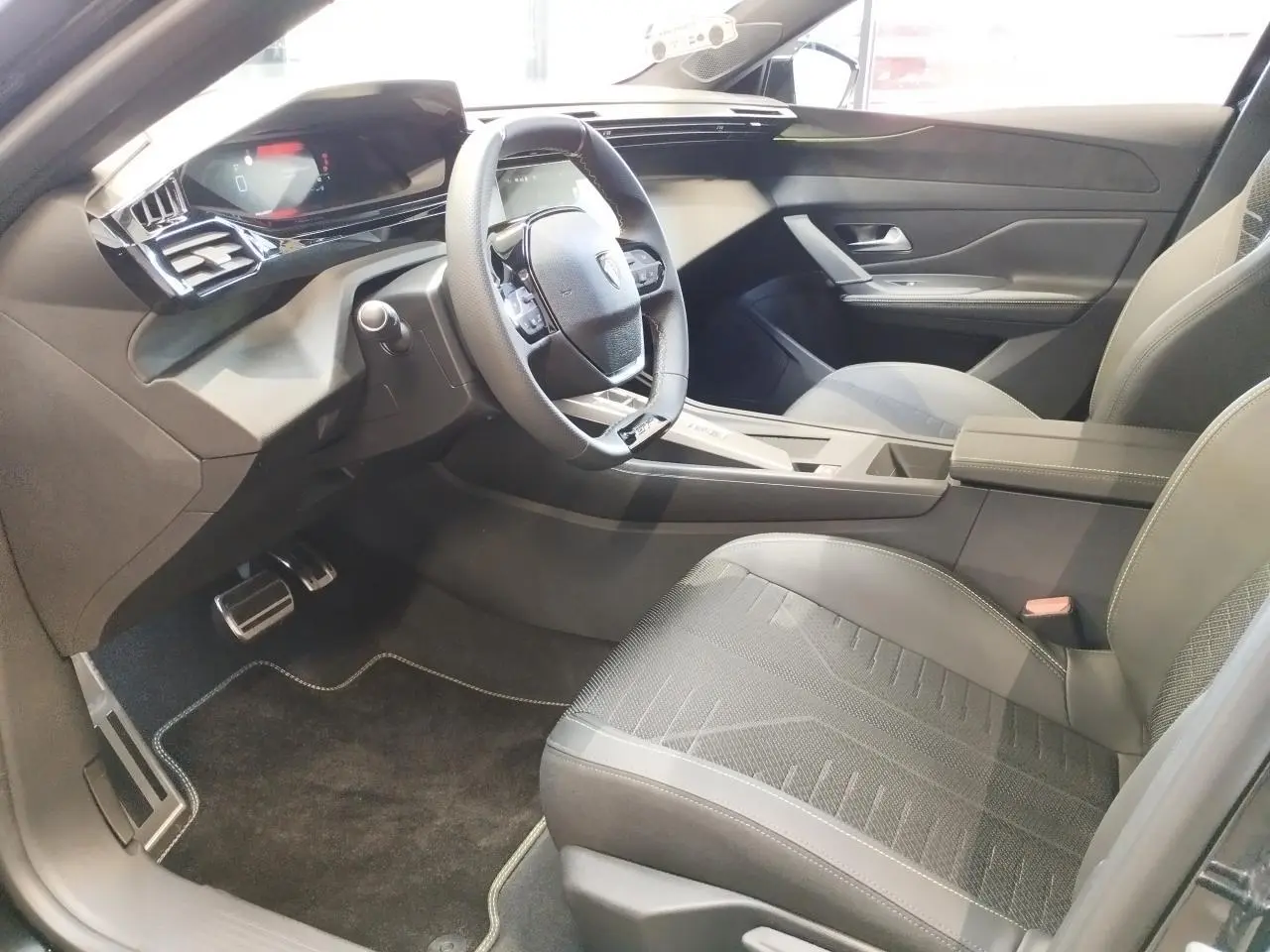 Vue intérieure côté conducteur de la Peugeot 308 hybride 2026, avec volant compact GT et sellerie noire détaillée.