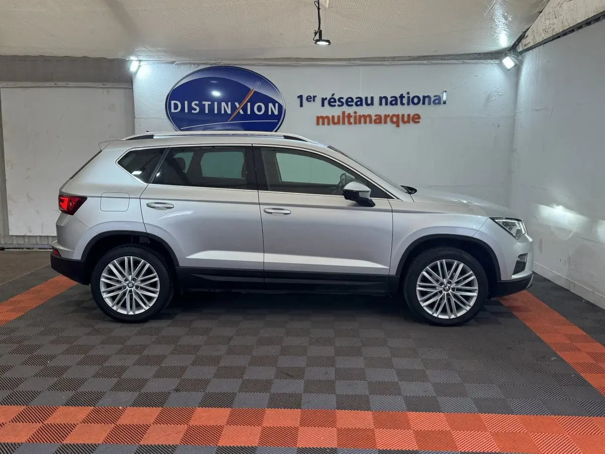 Profil côté droit d'un SUV SEAT Ateca gris clair 2019 avec jantes alliage et barres de toit chromées en intérieur showroom.