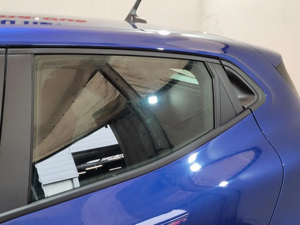 Détail de la vitre arrière côté gauche d'une Renault Clio bleu 2025 avec carrosserie lisse et reflet d'atelier visible.