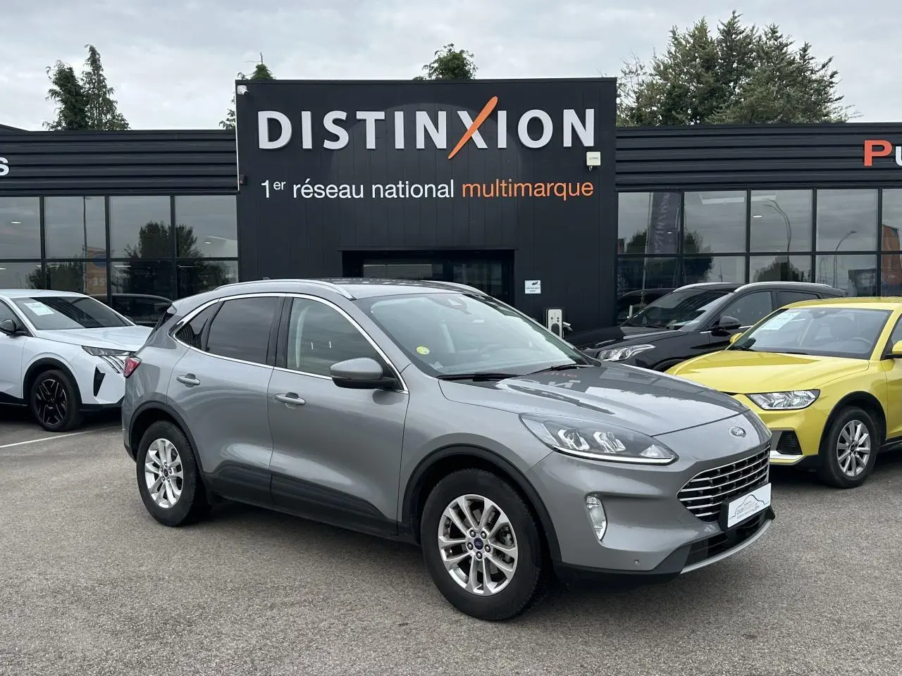 Ford Kuga gris solaire vu en 3/4 avant droit avec barres de toit aluminium sur parking devant un bâtiment Distinxion.