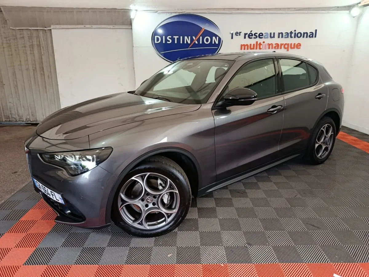 Alfa Romeo Stelvio gris foncé vu de 3/4 avant droit, avec jantes distinctives et phares LED allumés.