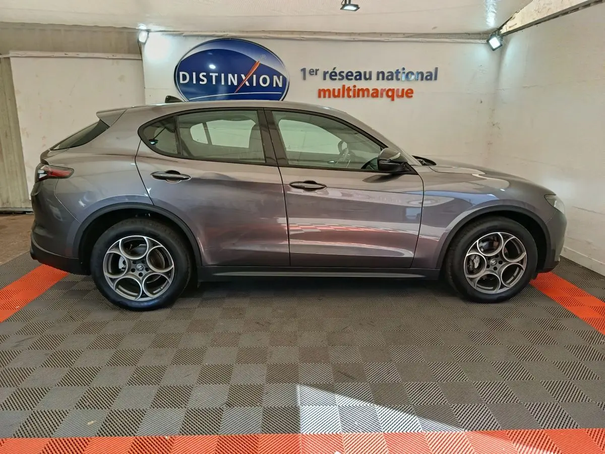Profil côté gauche de l'Alfa Romeo Stelvio gris foncé 2024 dans un showroom avec jantes spécifiques et logo Distinxion en arrière-plan.