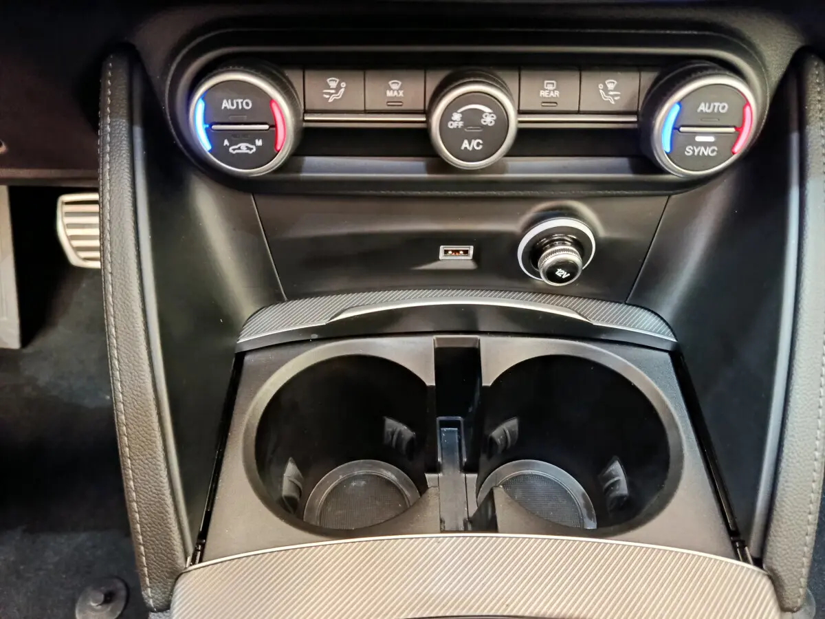 Gros plan sur la console centrale noire de l'Alfa Romeo Stelvio 2024 avec commandes climatisation et prises USB et 12V.