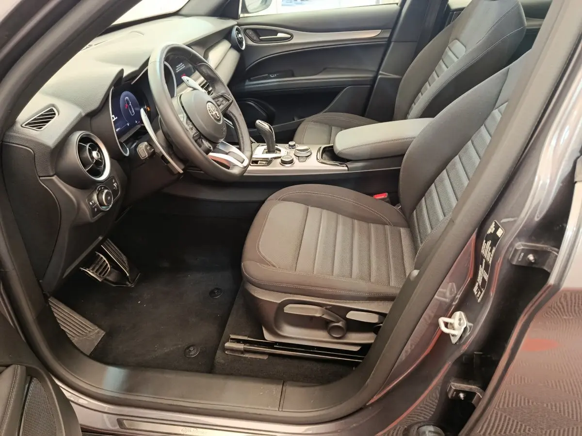 Vue côté conducteur intérieur de l'Alfa Romeo Stelvio gris foncé 2024, avec sièges tissu et tableau de bord moderne.
