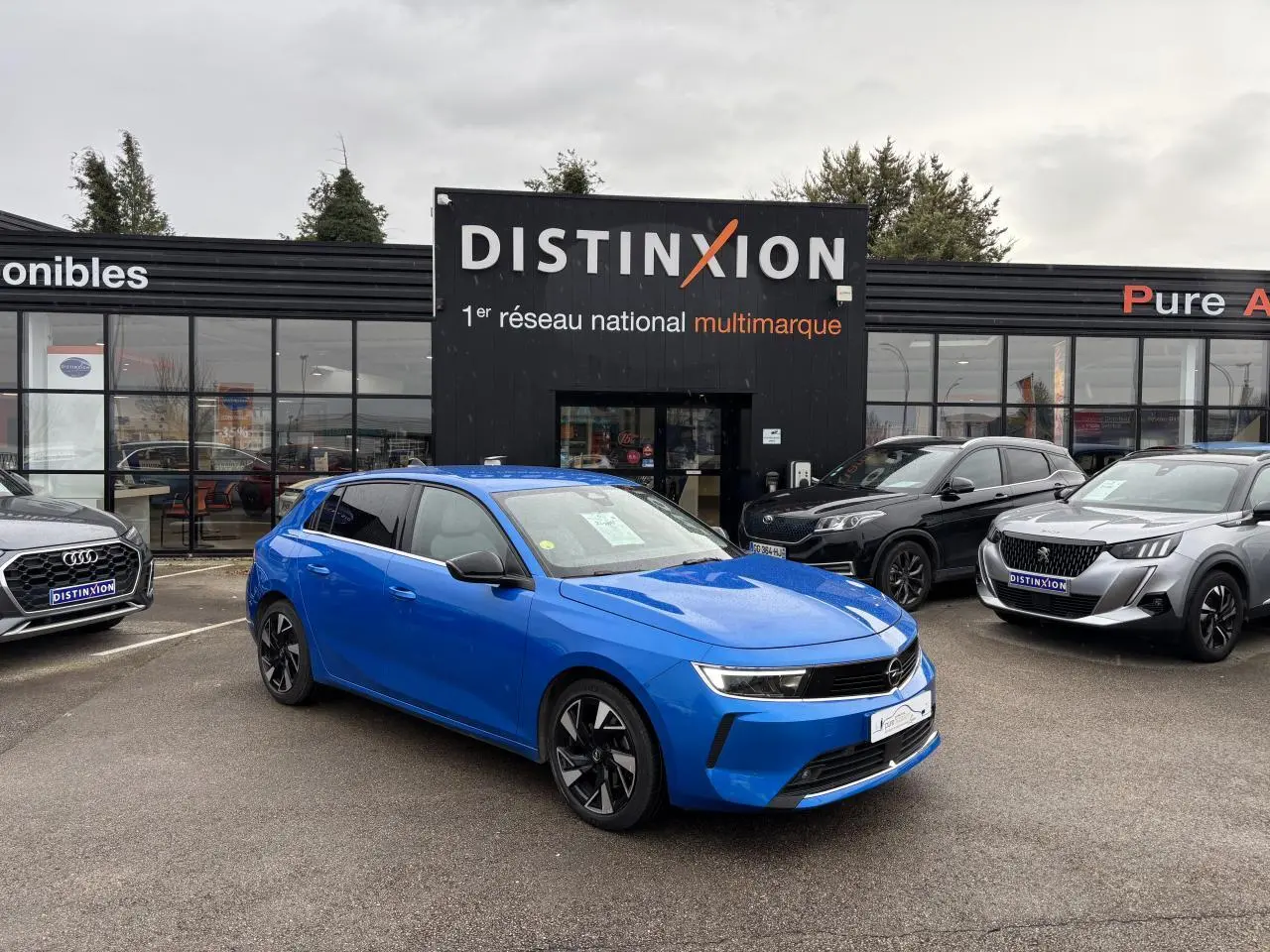 Opel Astra VI bleu clair vue 3/4 avant droit avec jantes noires devant concession Distinxion