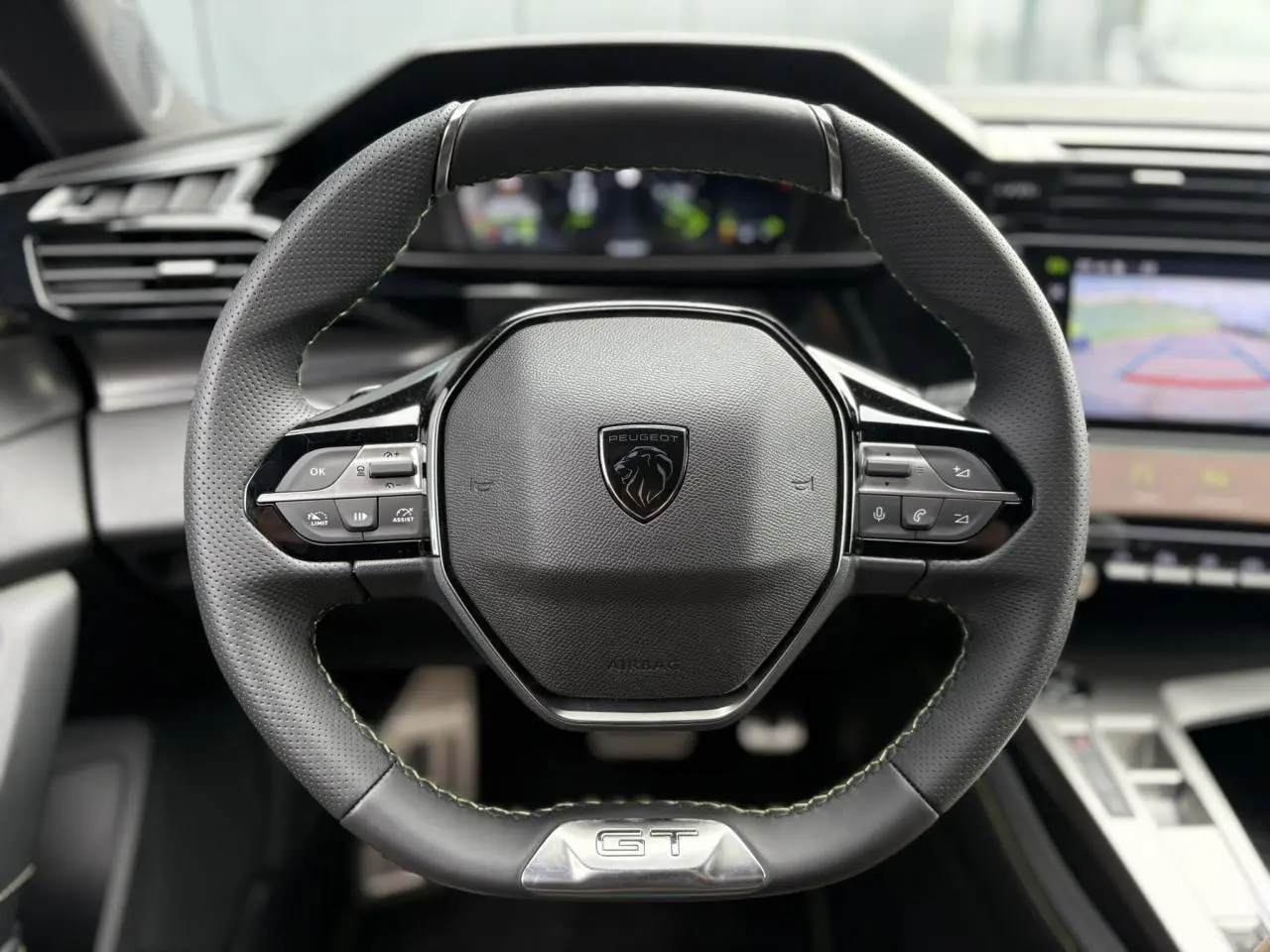 Volant cuir noir Peugeot 308 GT avec logo lion et badge GT, tableau de bord numérique et écran de caméra de recul visibles.
