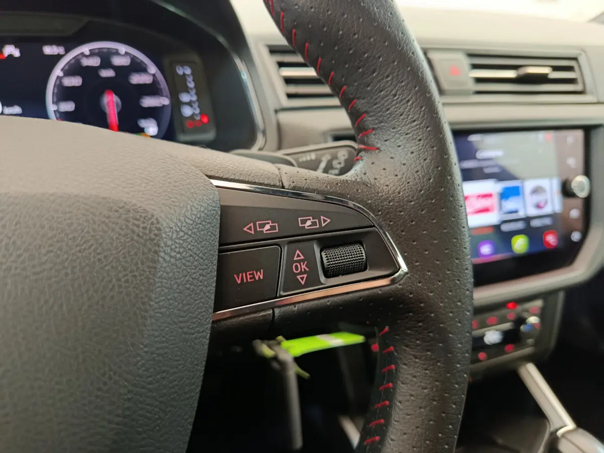 Gros plan sur le volant cuir noir avec surpiqûres rouges du SEAT Arona 2021, tableau de bord et écran tactile en arrière-plan.