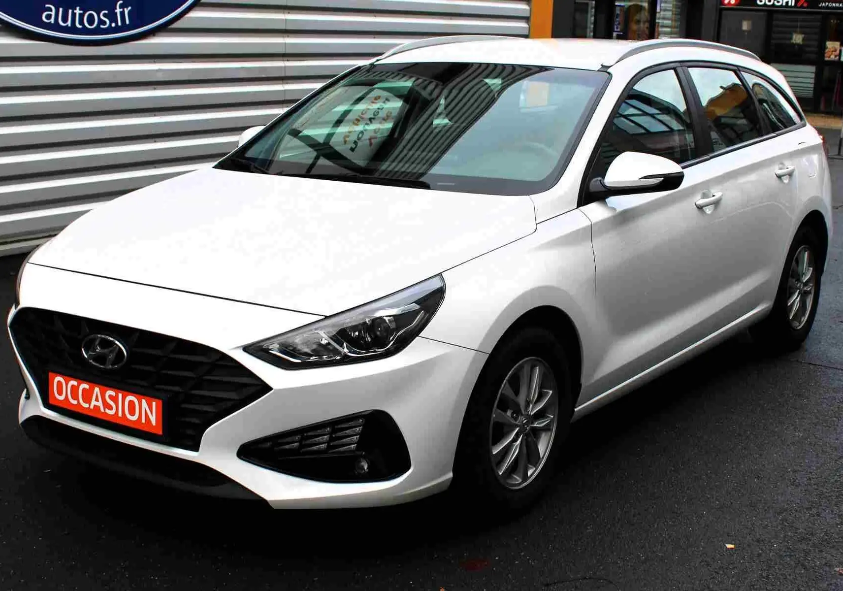 Hyundai i30 SW blanc polaire vue 3/4 avant droit, avec calandre noire et plaque "OCCASION" rouge visible.