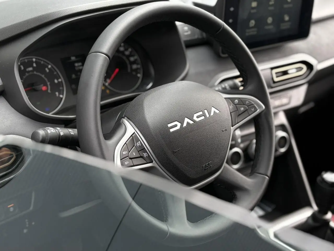 Vue rapprochée du volant DACIA noir avec commandes intégrées et tableau de bord du Jogger 2025 en arrière-plan.