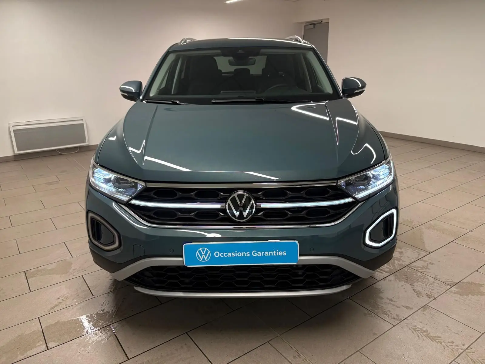 Vue frontale d'un Volkswagen T-Roc 1.5 TSI bleu pétrole avec éclairage LED et calandre chromée dans un showroom.
