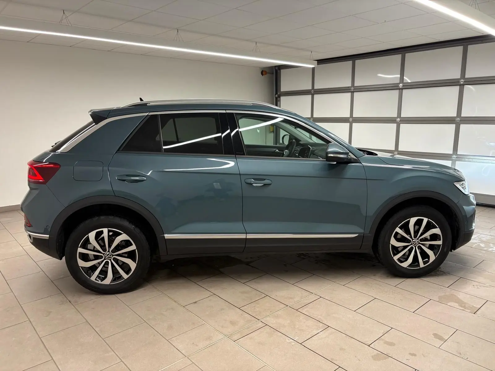 Vue de profil côté gauche d'un Volkswagen T-Roc bleu pétrole 2025 dans un garage, roues alliage visibles.