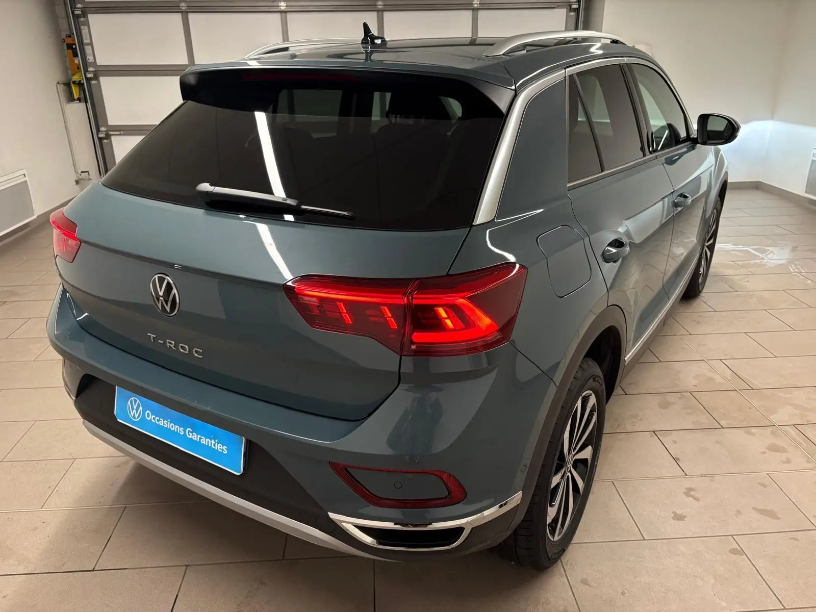 Vue 3/4 arrière droite du Volkswagen T-Roc bleu pétrole 2025 avec feux arrière allumés dans un garage.