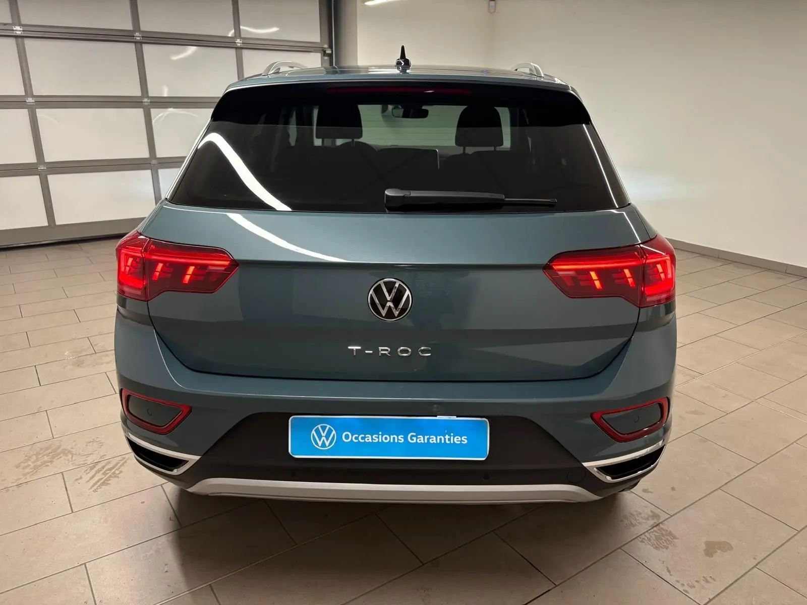 Vue arrière d'un Volkswagen T-Roc bleu pétrole 2025 dans un garage, avec feux arrière allumés et plaque "Occasions Garantis".