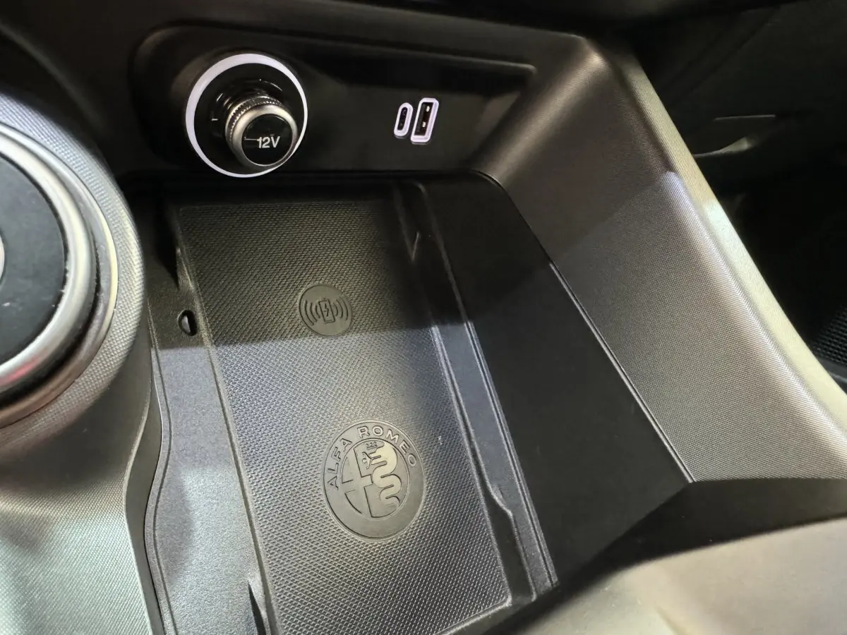 Gros plan sur la console centrale noire de l'Alfa Romeo Tonale rouge avec prise 12V et logo Alfa Romeo gravé.