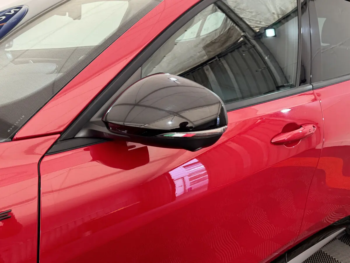 Gros plan sur le rétroviseur noir côté gauche d'une Alfa Romeo Tonale rouge brillante, avec poignée de porte visible.