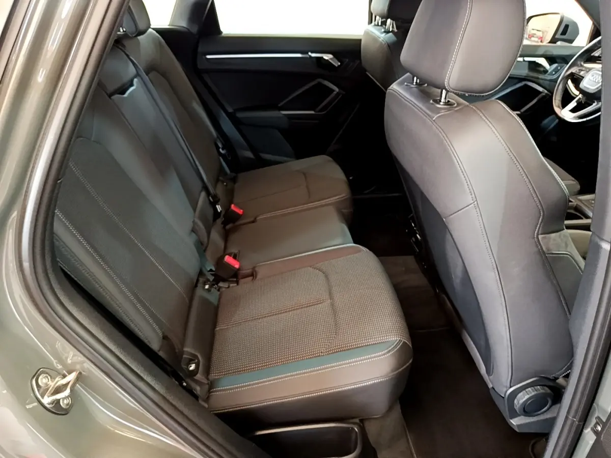 Vue intérieure côté arrière gauche de l’Audi Q3 gris foncé, montrant la banquette arrière en tissu et cuir avec ceintures rouges.