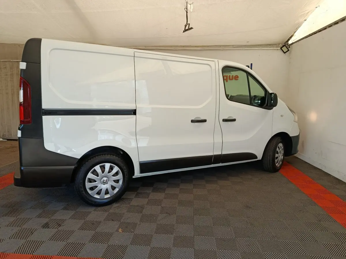Profil droit d’un utilitaire Renault Trafic blanc avec cloison pleine, stationné sur sol à damier gris et rouge.