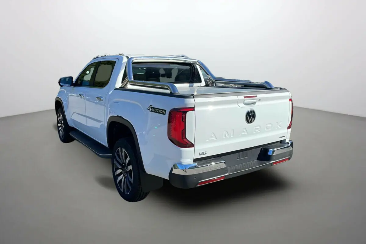Volkswagen Amarok V6 blanc vu de l'arrière côté gauche, avec badge 4MOTION et benne équipée d'arceaux chromés.
