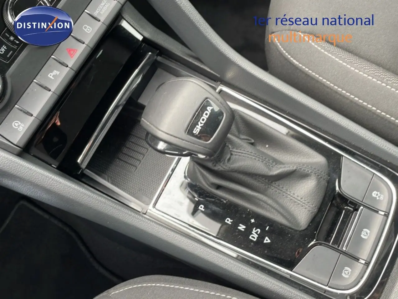 Gros plan sur la commande de boîte automatique noire avec logo SKODA, entourée de la console centrale et siège tissu gris.
