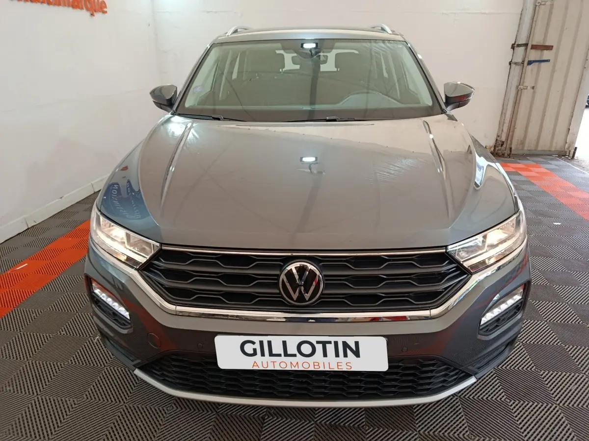 Vue avant d'un Volkswagen T-Roc gris foncé avec calandre hexagonale et phares LED allumés dans un showroom.