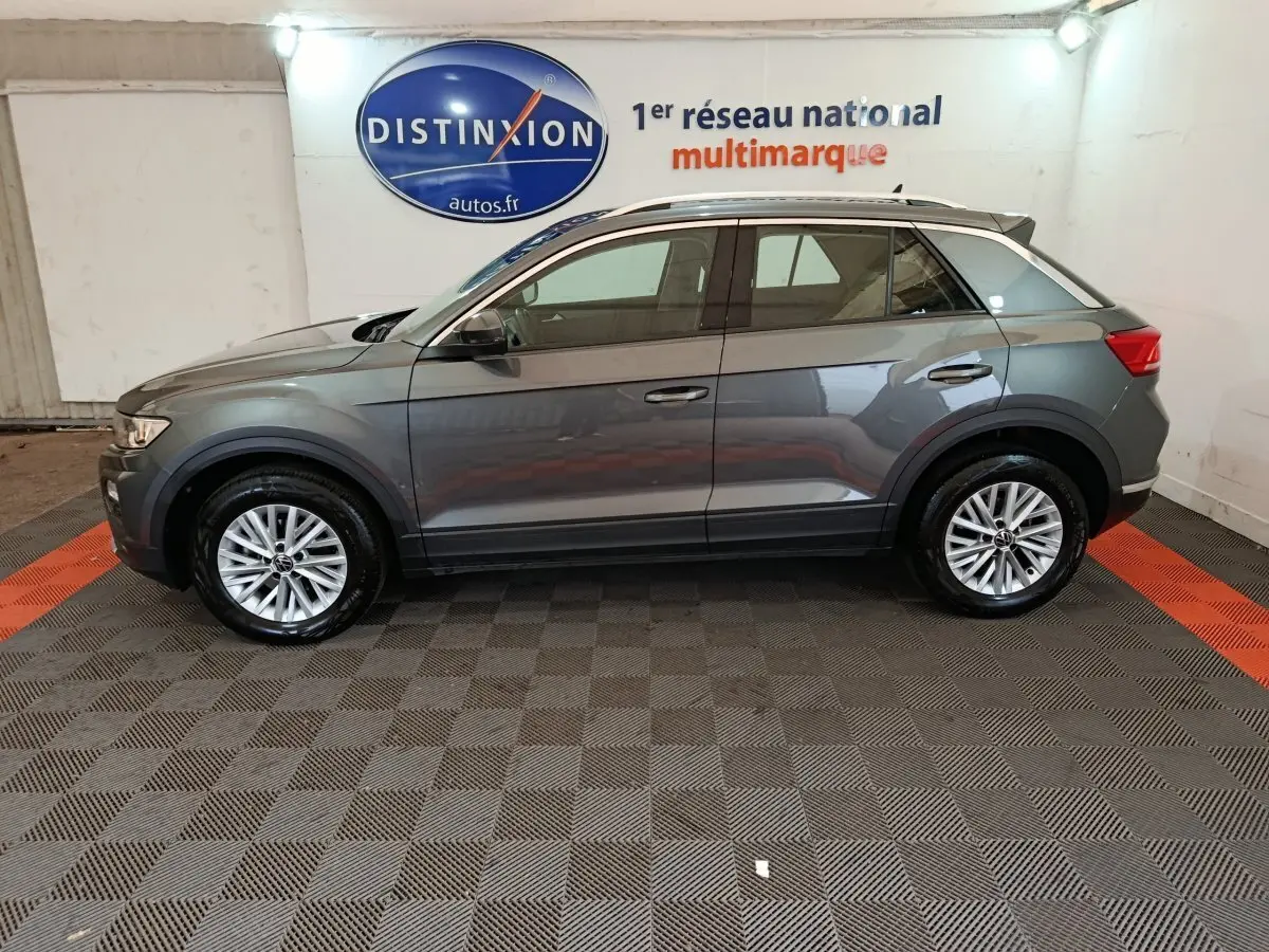 Profil côté gauche d'un Volkswagen T-Roc gris foncé 2021 dans un showroom avec sol damier et logo Distinxion en arrière-plan