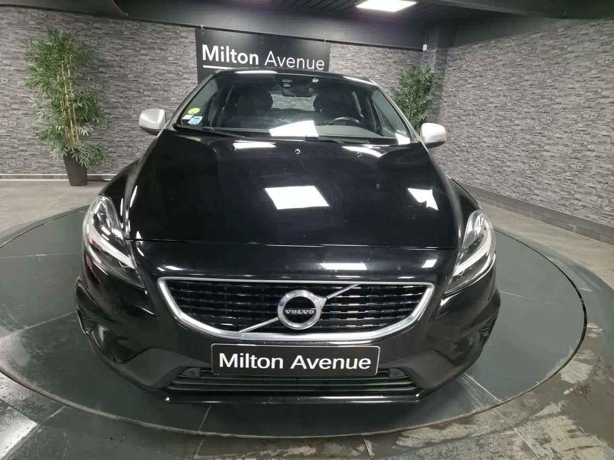 Vue avant d'une Volvo V40 D2 R-Design noire avec calandre chromée et plaque "Milton Avenue" en intérieur showroom.