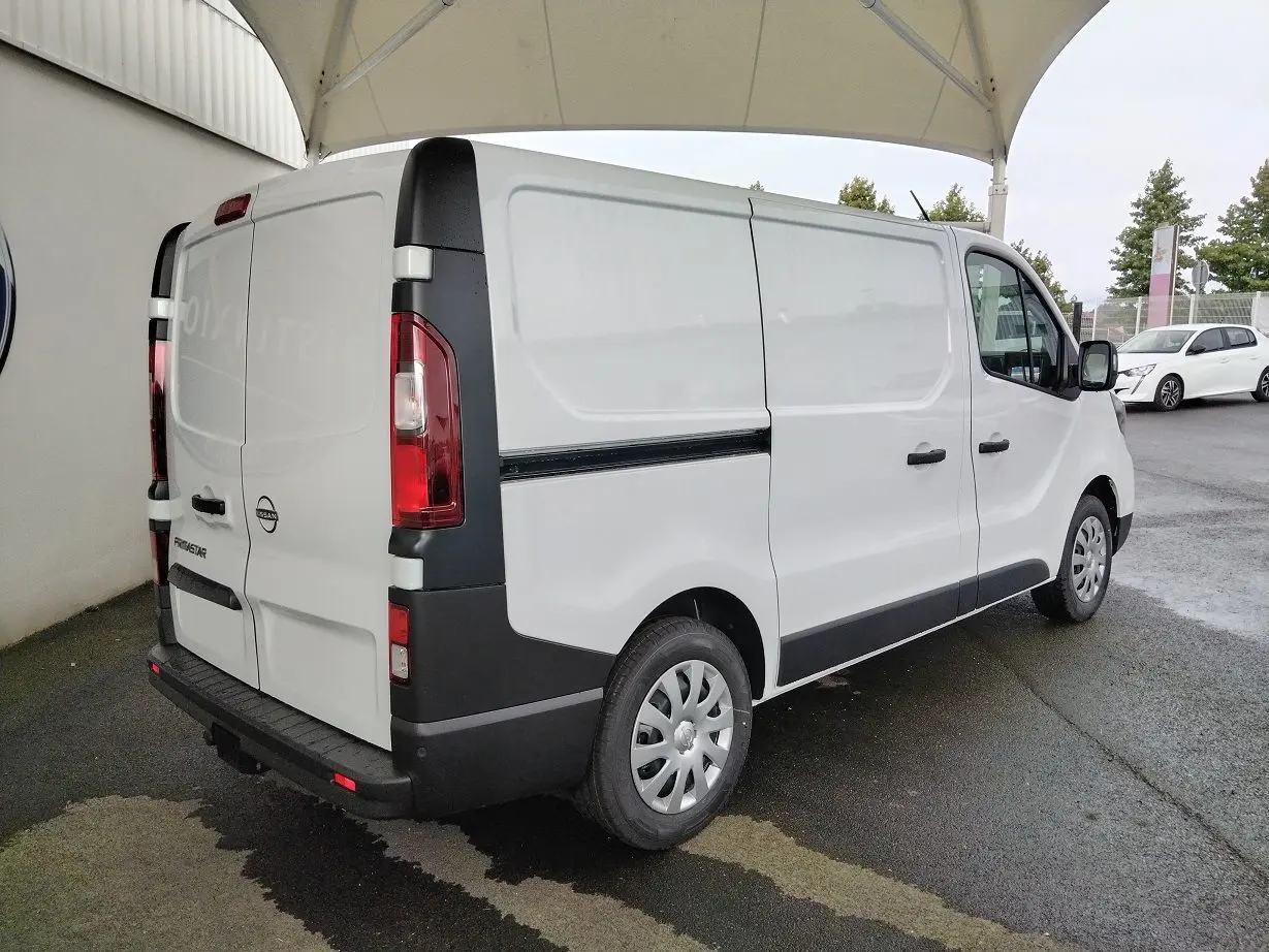 Vue 3/4 arrière droite d'un utilitaire Nissan Primastar blanc avec attelage et protections noires sur pare-chocs et bas de caisse.