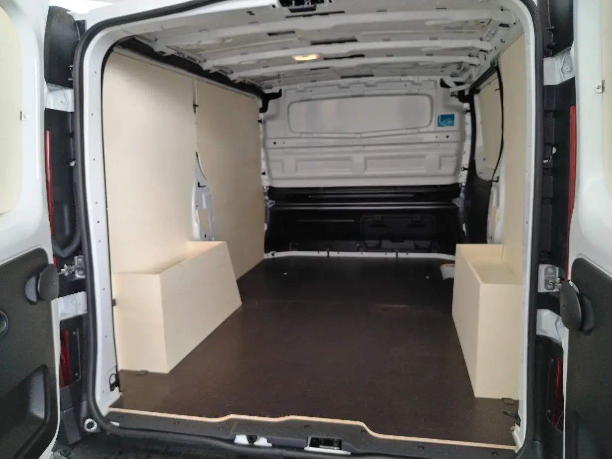 Vue intérieure arrière du fourgon blanc Nissan Primastar 2025 avec aménagement bois et plancher noir.