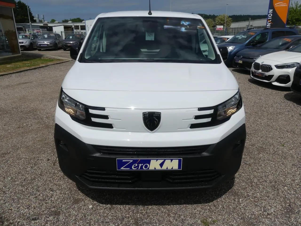 Vue frontale d'un Peugeot Partner blanc avec calandre noire et logo lion au centre, sur parking extérieur.