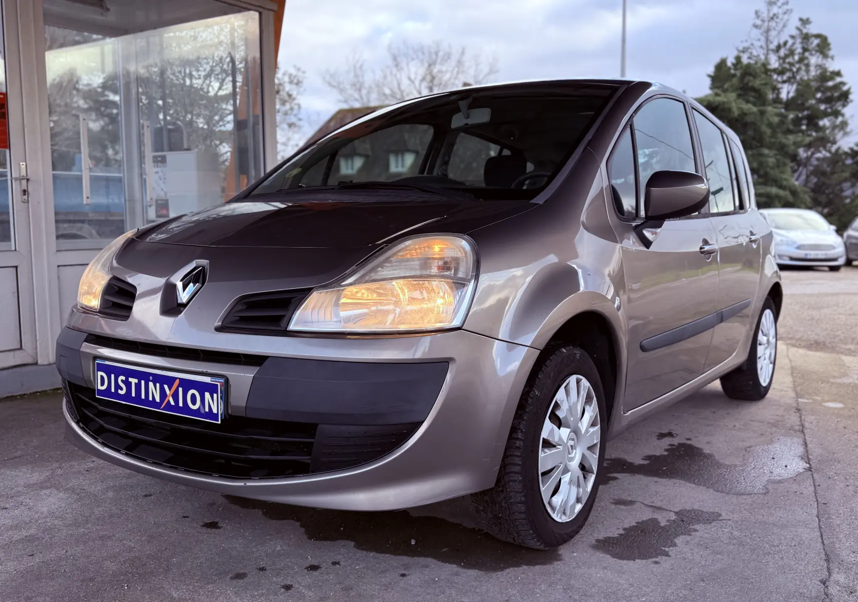 Renault Modus beige vue 3/4 avant droit avec phares allumés et plaque Distinxion visible.