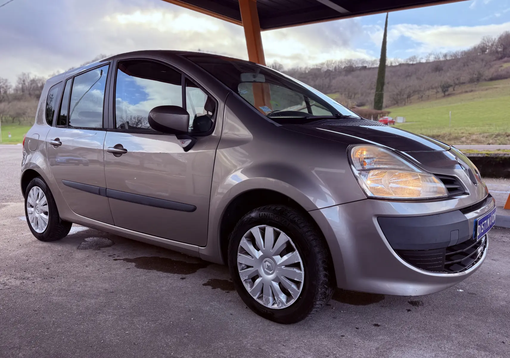 Renault Modus beige vue 3/4 avant droit sous abri, phare avant droit allumé et jantes avec enjoliveurs.