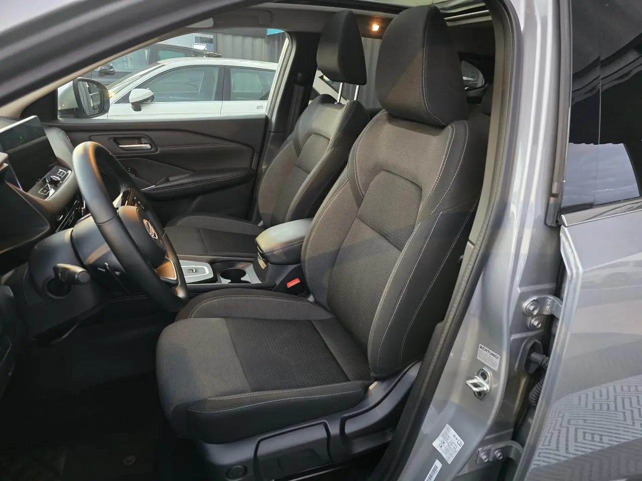 Intérieur avant du Nissan Qashqai gris silver 2024, sièges tissu noirs et accoudoir central visible côté conducteur.