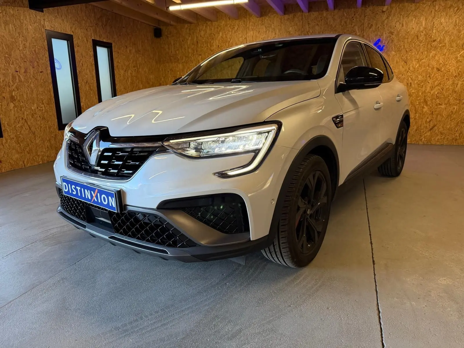 Vue avant d'un Renault Arkana blanc nacré avec calandre noire et phares LED allumés en intérieur showroom.