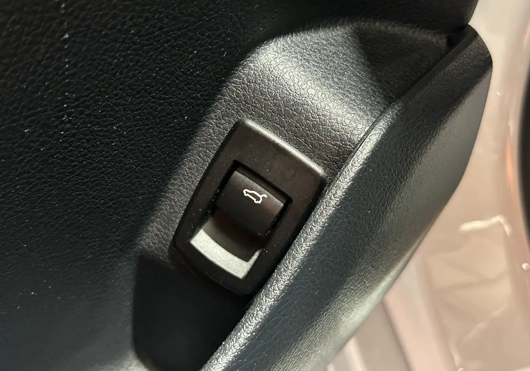 Gros plan sur le bouton d'ouverture du coffre, entouré de plastique noir, avec une partie de la carrosserie blanche visible en arrière-plan.