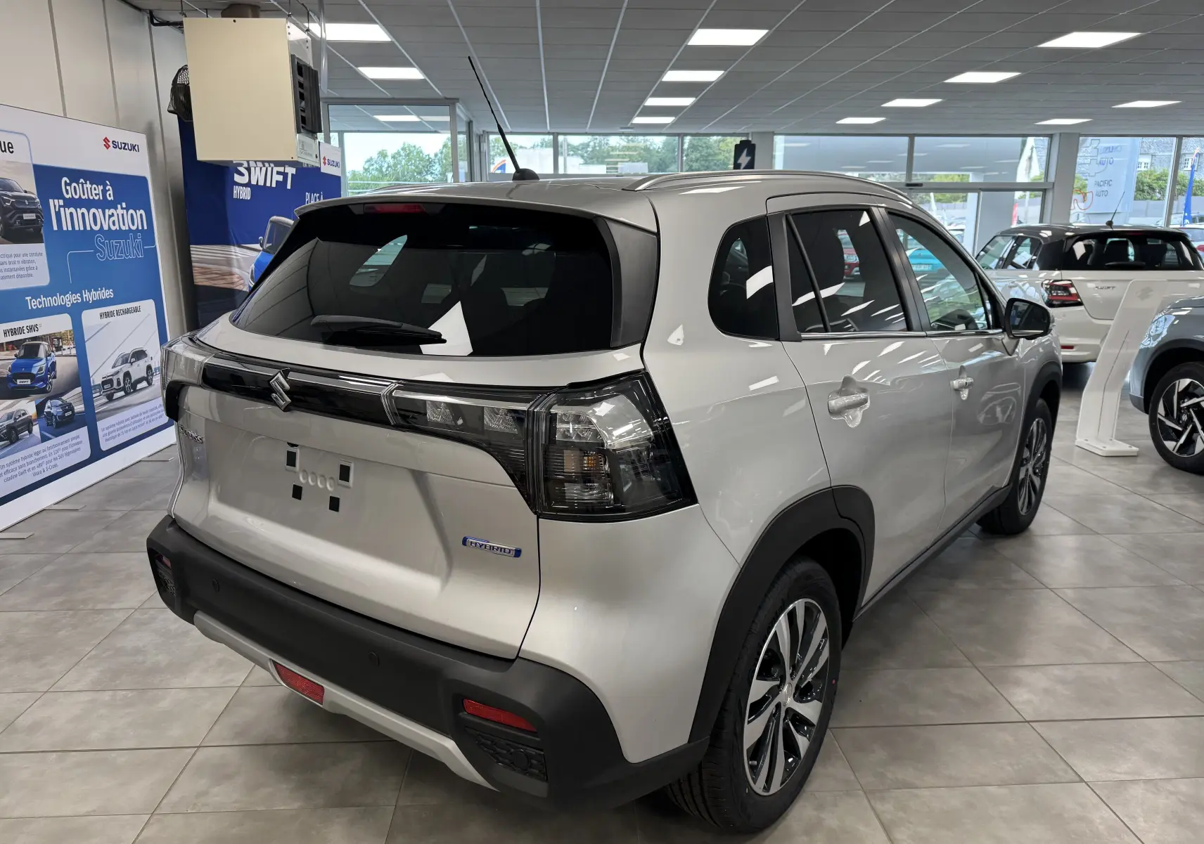 Vue 3/4 arrière droite d'un Suzuki S-Cross gris hybride 2025 en showroom, avec feux arrière fumés et jantes alliage.