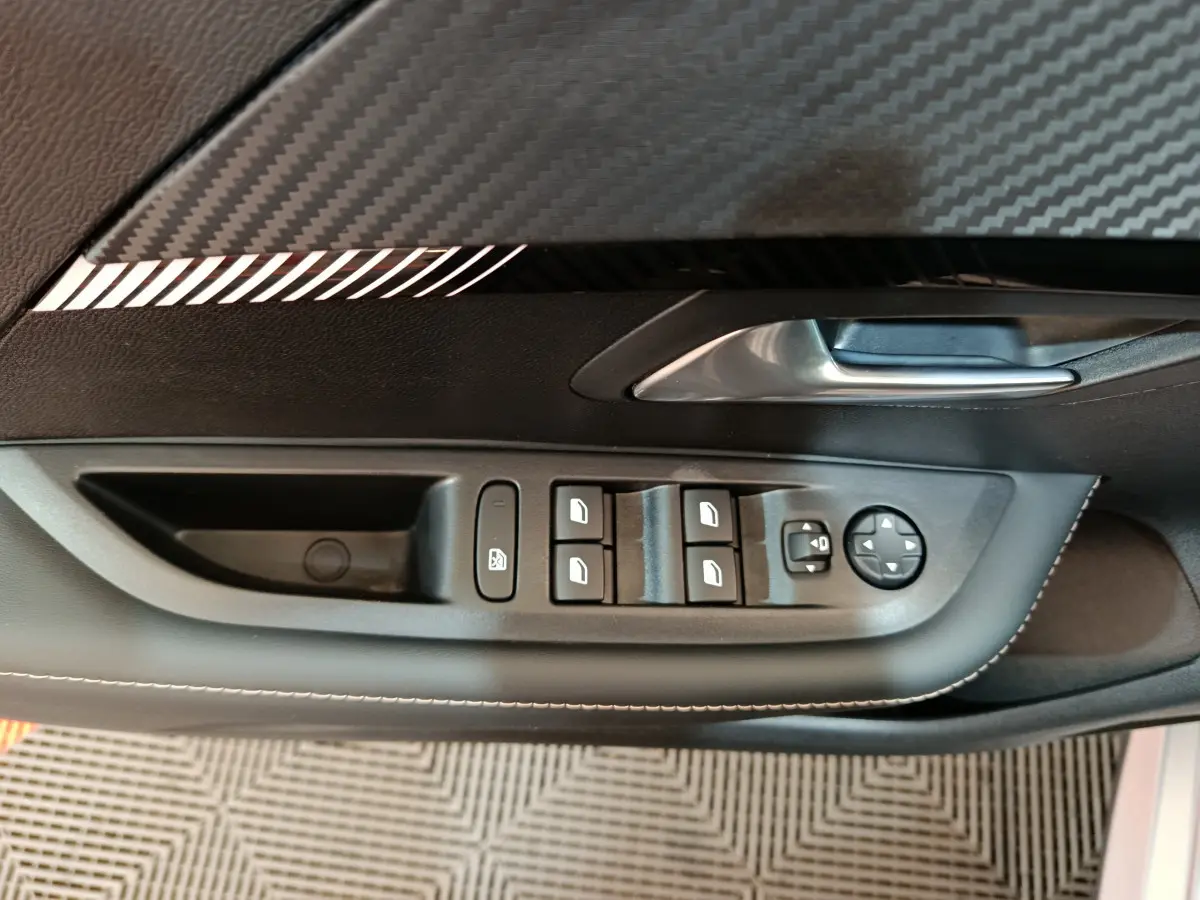 Gros plan sur la commande des vitres et rétroviseurs côté conducteur d'une Peugeot 208 gris clair, avec surpiqûres blanches.