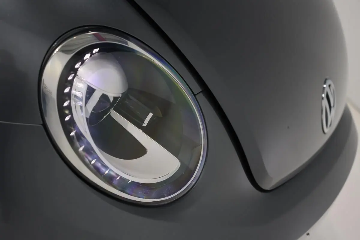 Gros plan sur le phare avant gauche gris d’une Volkswagen Coccinelle 2016 avec logo VW visible sur le capot.