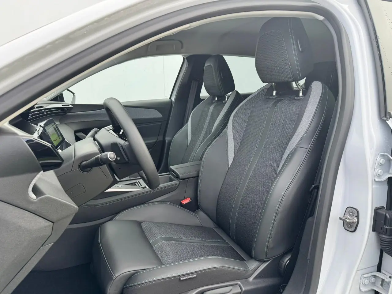 Vue intérieure côté conducteur de la Peugeot 308 SW 2024 blanc Okenite avec sièges tri matières noirs et gris à surpiqûres bleues.