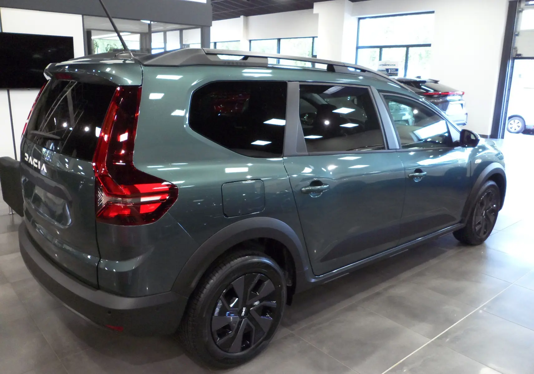 Vue 3/4 arrière droite d'un Dacia Jogger vert cèdre avec feux arrière en forme de C et barres de toit noires.