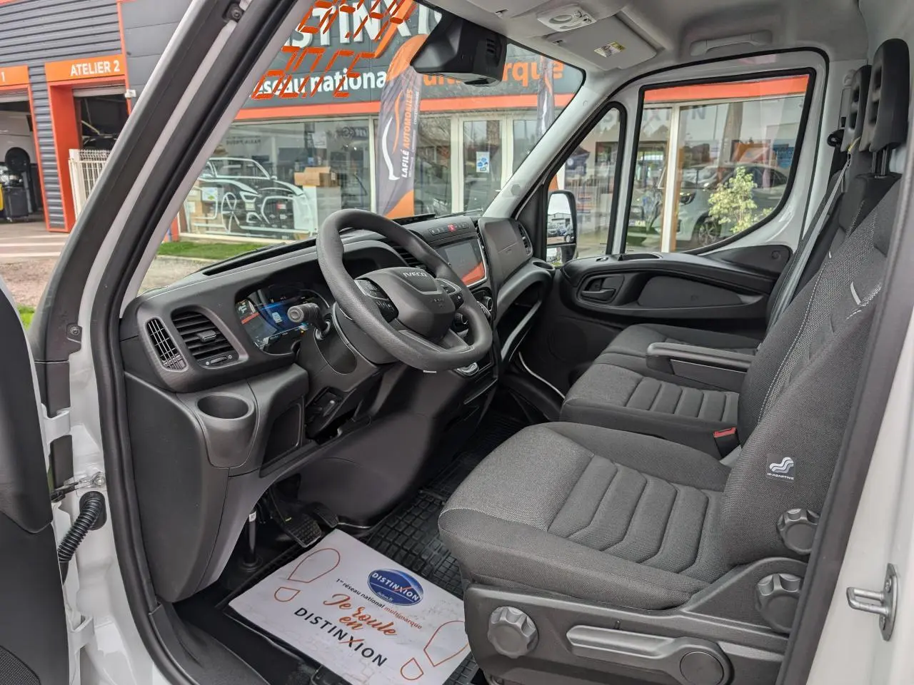 Intérieur du Iveco Daily blanc 2025 vu côté conducteur, montrant le tableau de bord et les sièges en tissu gris.