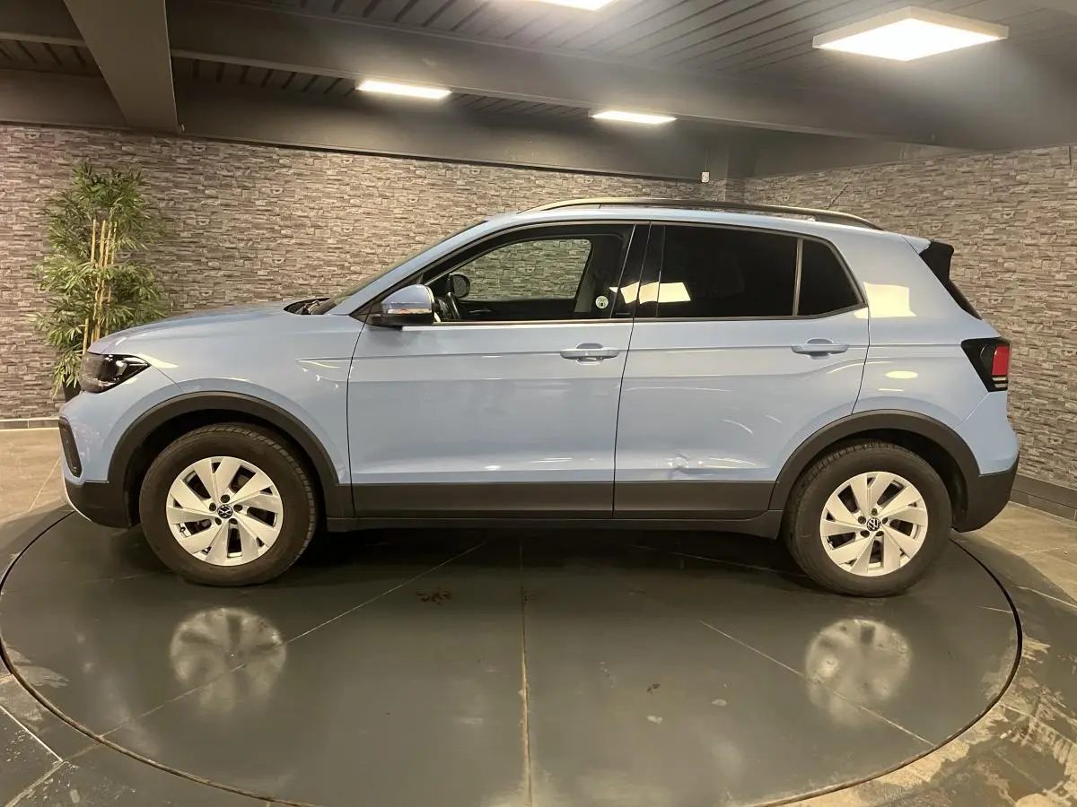 Profil côté gauche du Volkswagen T-Cross bleu clair 2024 avec vitres arrière surteintées en intérieur showroom.
