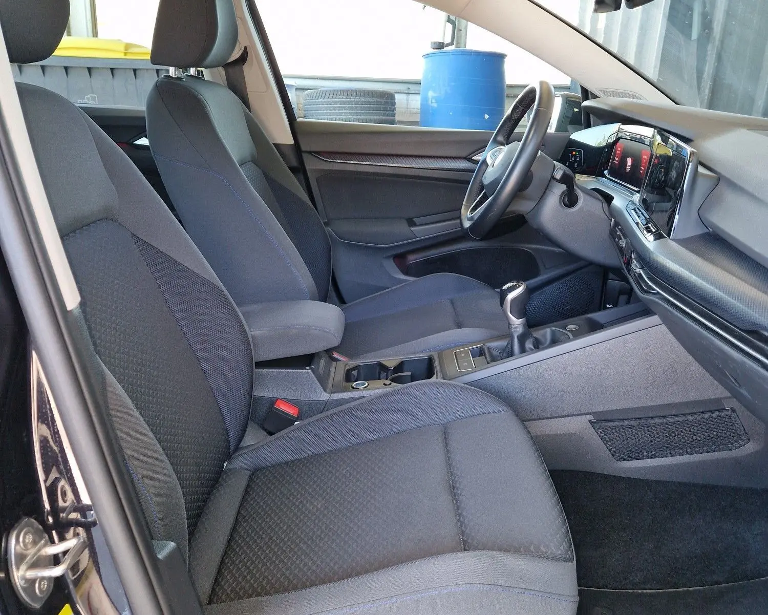 Intérieur côté conducteur d'une Volkswagen Golf VIII noire, sièges tissu gris et tableau de bord moderne avec boîte manuelle.