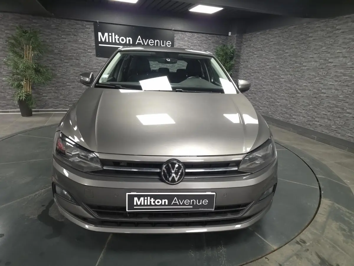Volkswagen Polo gris métallisé vue de face en intérieur avec logo VW et plaque Milton Avenue visible.