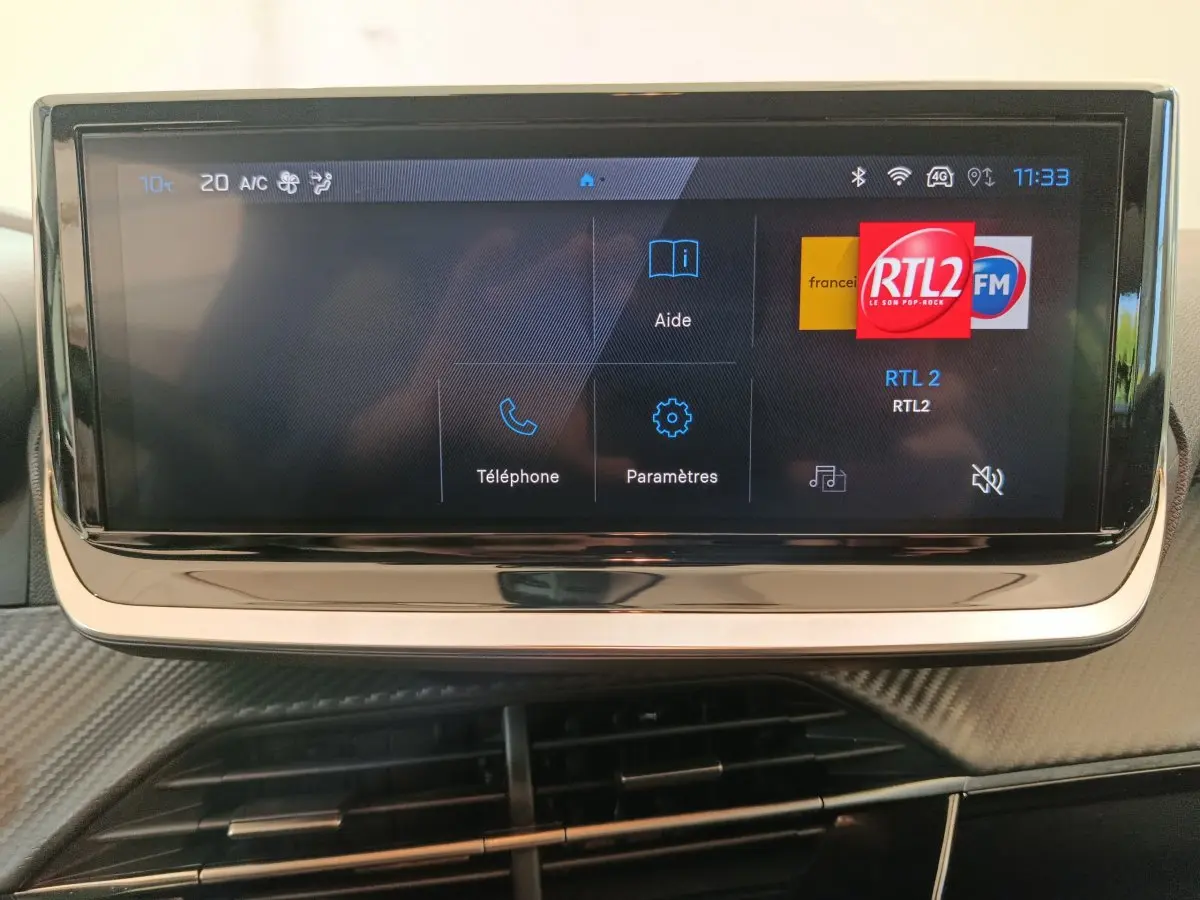 Écran tactile central affichant la radio RTL2 dans l'habitacle d'une Peugeot 208 gris foncé, vue frontale rapprochée.