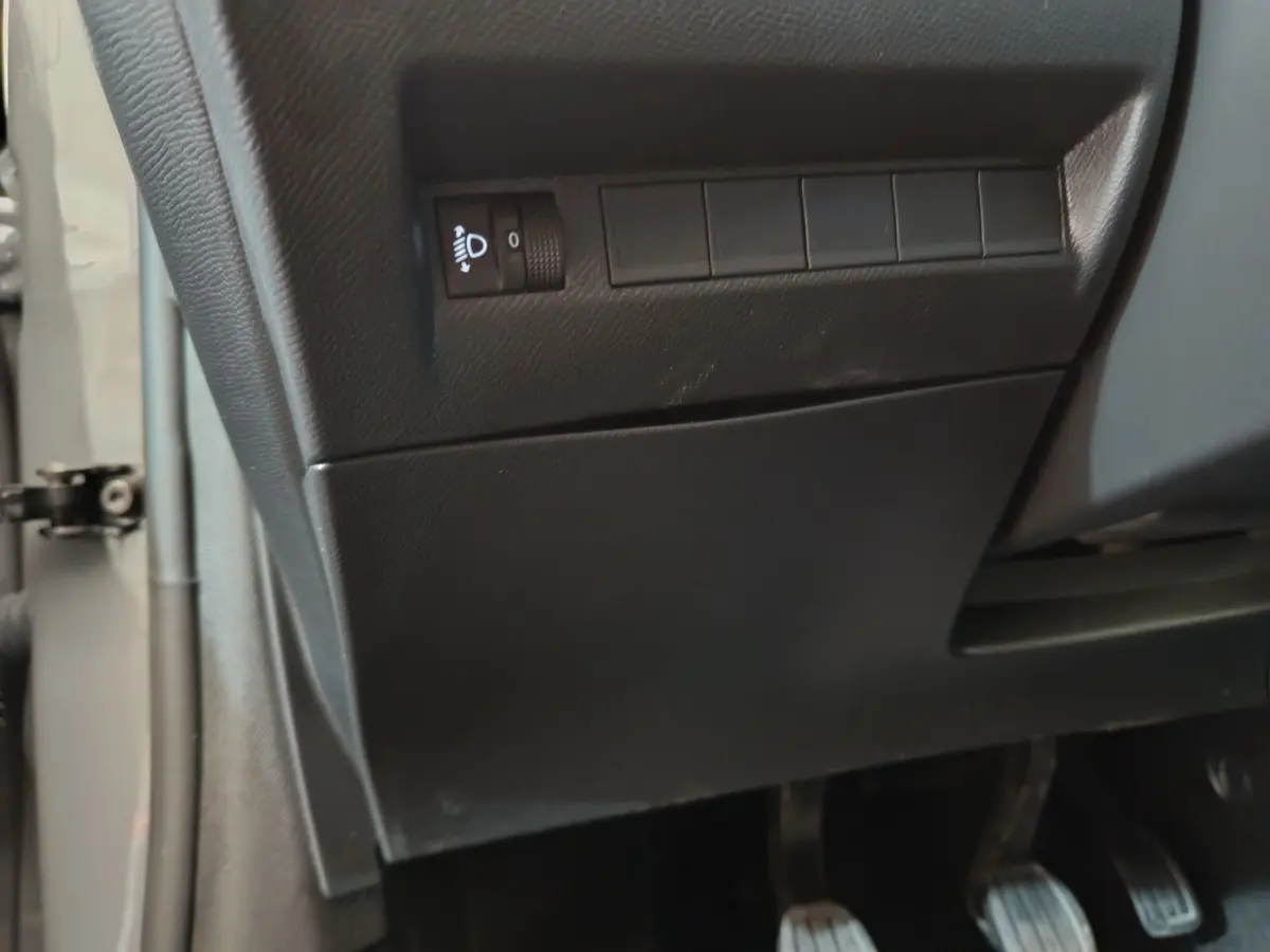 Détail du tableau de bord côté conducteur de la Peugeot 208 gris foncé, avec commande réglage hauteur phares et pédales visibles.